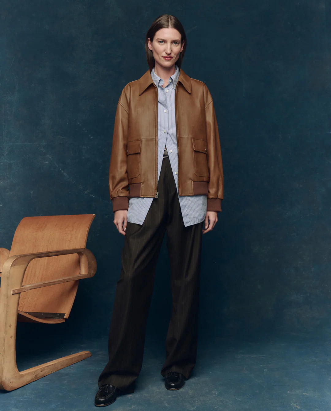 A person stands indoors wearing The Flight Jacket by The Great Inc. over a blue button-up shirt and loose dark trousers with black shoes. A wooden chair sits to the left, set against a dark blue background.
