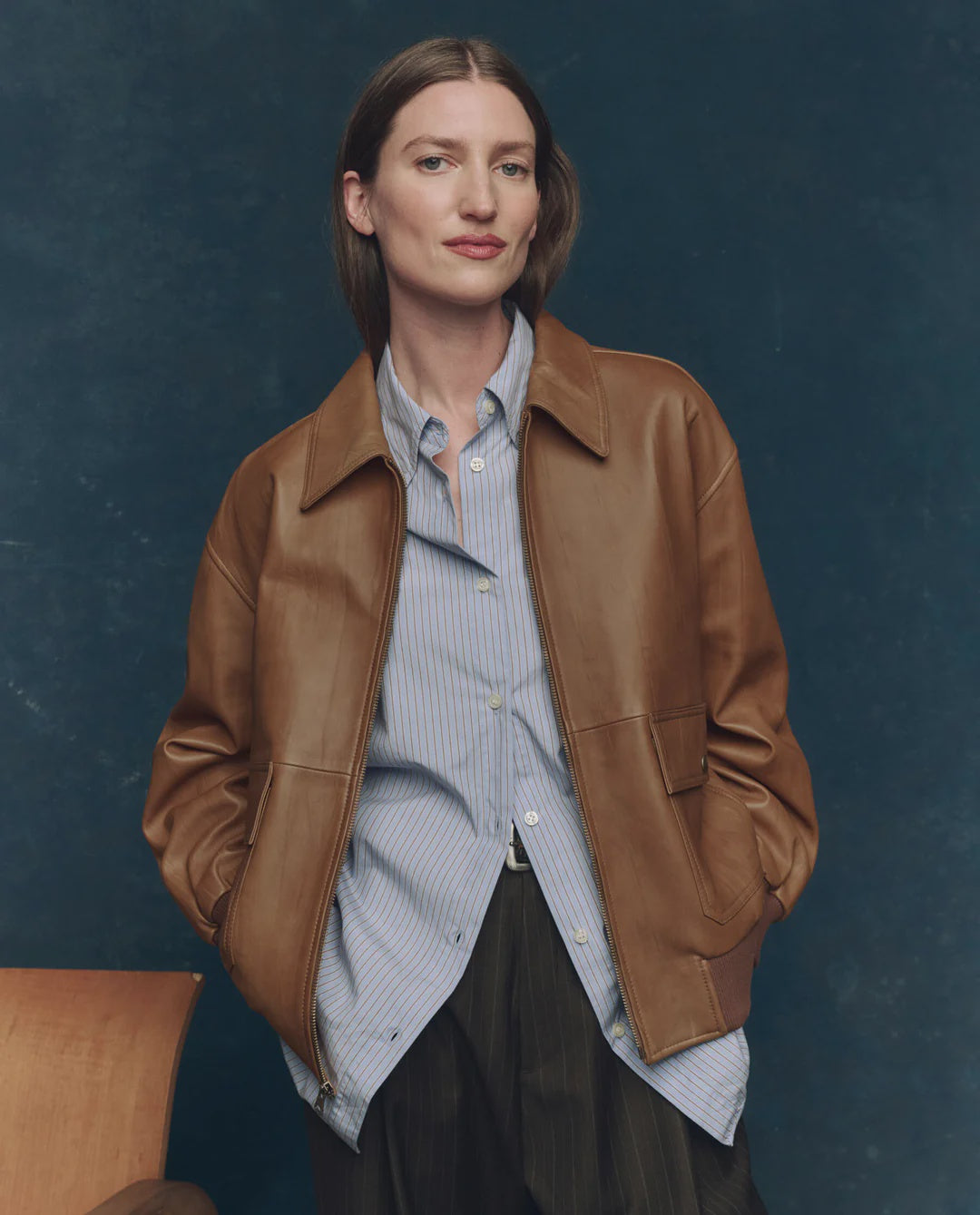 A woman with straight brown hair wears The Great Inc.’s vintage-inspired Flight Jacket over a striped button-up shirt and black pants, standing with her hands in her jacket pockets against a dark blue background.