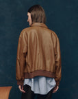 A person with shoulder-length brown hair faces away, wearing The Flight Jacket by The Great Inc. over a long blue striped shirt. The dark blue background features a partially visible wooden chair on the left.