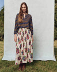 Standing outdoors on grass with green trees, a person wears The Great Inc.'s Viola Skirt, a voluminous floral piece with a smocked waistband, and a shadow stripe cotton long-sleeve shirt.