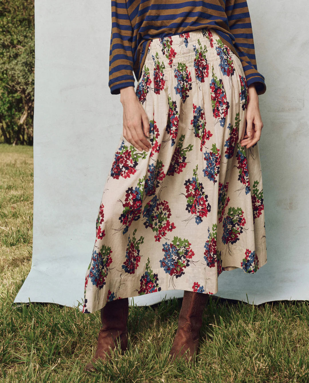 A person in a shadow stripe cotton long-sleeve top and The Viola Skirt by The Great Inc. stands outdoors. The cream-colored skirt, featuring red and blue flowers with a smocked waistband, is paired with brown boots against a grassy backdrop.