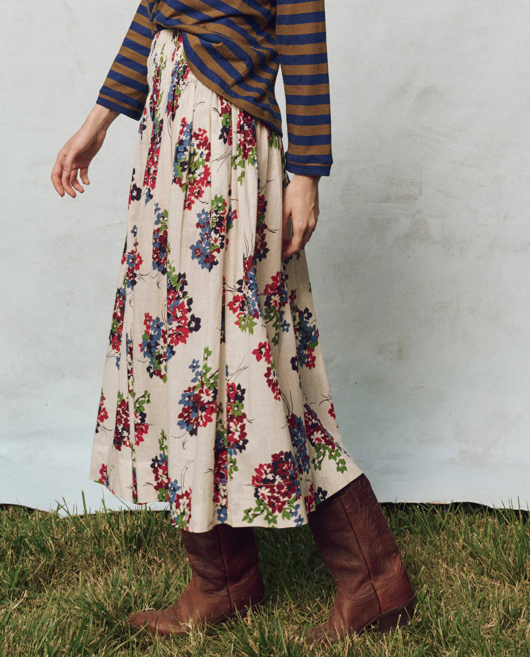 A person wears a striped long-sleeve shirt and The Great Inc.'s Viola Skirt with a smocked waistband on grass, paired with brown cowboy boots. Shadow stripe textured cotton adds interest to the neutral setting.