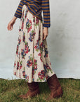 A person wears a striped long-sleeve shirt and The Great Inc.'s Viola Skirt with a smocked waistband on grass, paired with brown cowboy boots. Shadow stripe textured cotton adds interest to the neutral setting.