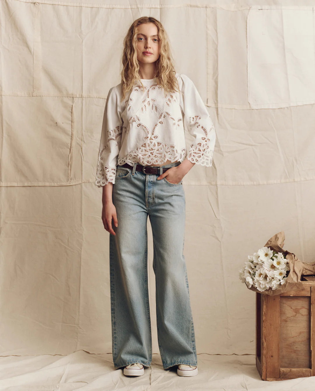 A woman stands before a neutral fabric backdrop, wearing The Picnic Top by The Great Inc., light-wash wide-leg jeans, and white sneakers. A wooden box with white flowers is positioned to her right.