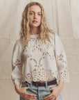 A woman with long, wavy blonde hair wears The Picnic Top by The Great Inc.—a white crochet lace long-sleeve blouse—paired with light blue jeans and a brown belt. She stands against a beige fabric backdrop, hands in pockets, looking at the camera.