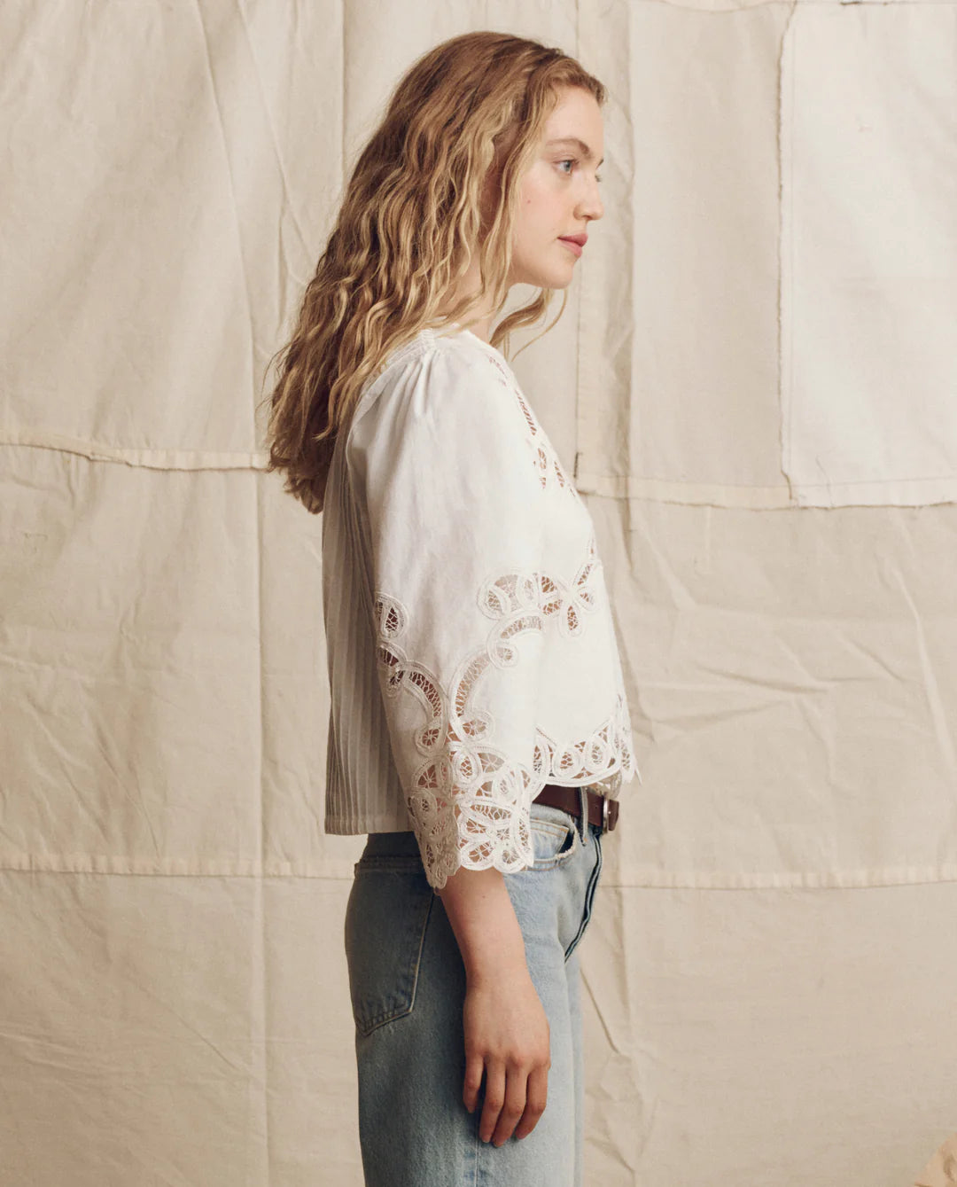 A woman with wavy blonde hair wears The Picnic Top by The Great Inc., featuring Battenberg lace sleeve details, paired with light blue jeans, standing in profile against a neutral quilted fabric background.