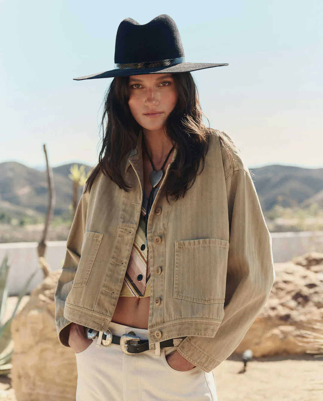A woman outdoors in a desert setting wears The Great Inc.'s The Rancher Jacket, styled with a wide-brimmed black hat, patterned shirt, and light pants amid mountains and cacti.