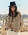 A woman outdoors in a desert setting wears The Great Inc.'s The Rancher Jacket, styled with a wide-brimmed black hat, patterned shirt, and light pants amid mountains and cacti.
