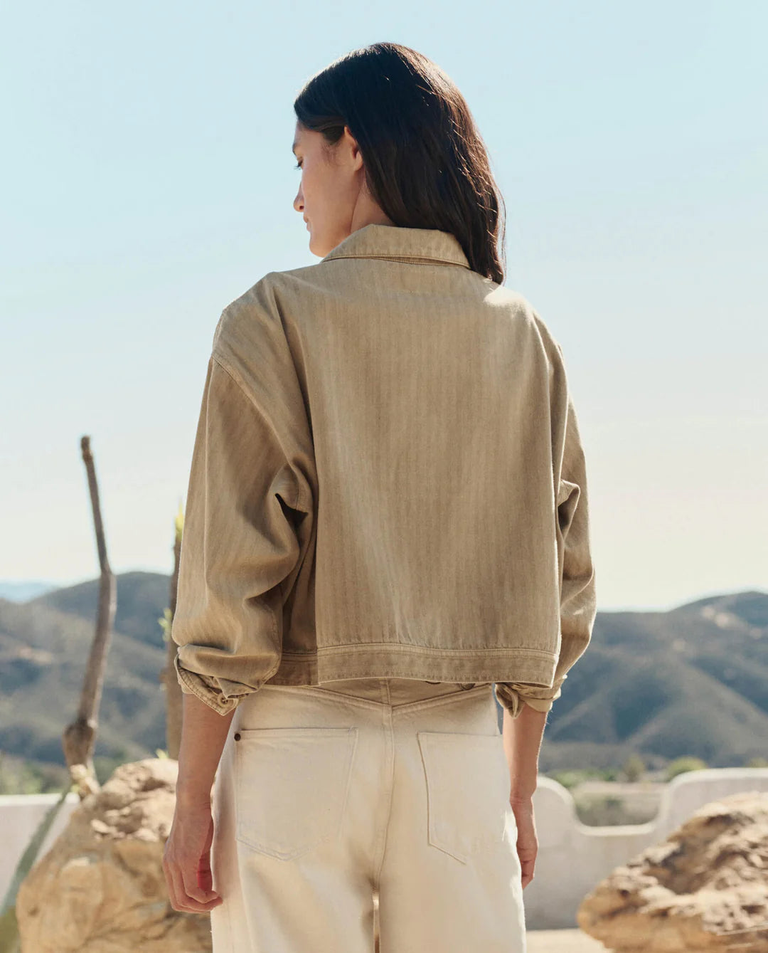 A person with long, dark hair stands outdoors facing away, wearing The Rancher Jacket by The Great Inc. with cream pants. Rocky hills and a clear sky are in the background.