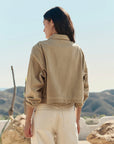 A person with long, dark hair stands outdoors facing away, wearing The Rancher Jacket by The Great Inc. with cream pants. Rocky hills and a clear sky are in the background.