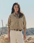 A woman with long dark hair stands outdoors in The Great Inc.'s The Rancher Jacket, beige cotton herringbone, paired with cream pants and a black belt. Rocky hills and a clear sky evoke an effortless vintage workwear look.