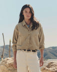 A woman with long dark hair stands outdoors in The Great Inc.'s The Rancher Jacket, beige cotton herringbone, paired with cream pants and a black belt. Rocky hills and a clear sky evoke an effortless vintage workwear look.