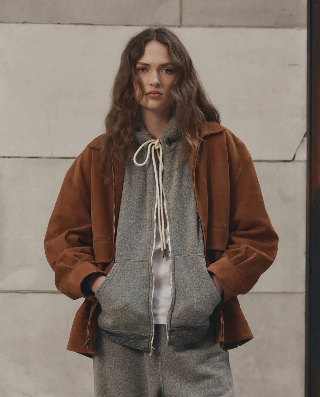 Brown fleece hoodie from The Great Inc. styled casually with sweatpants near a stone wall, relaxed lifestyle setting.
