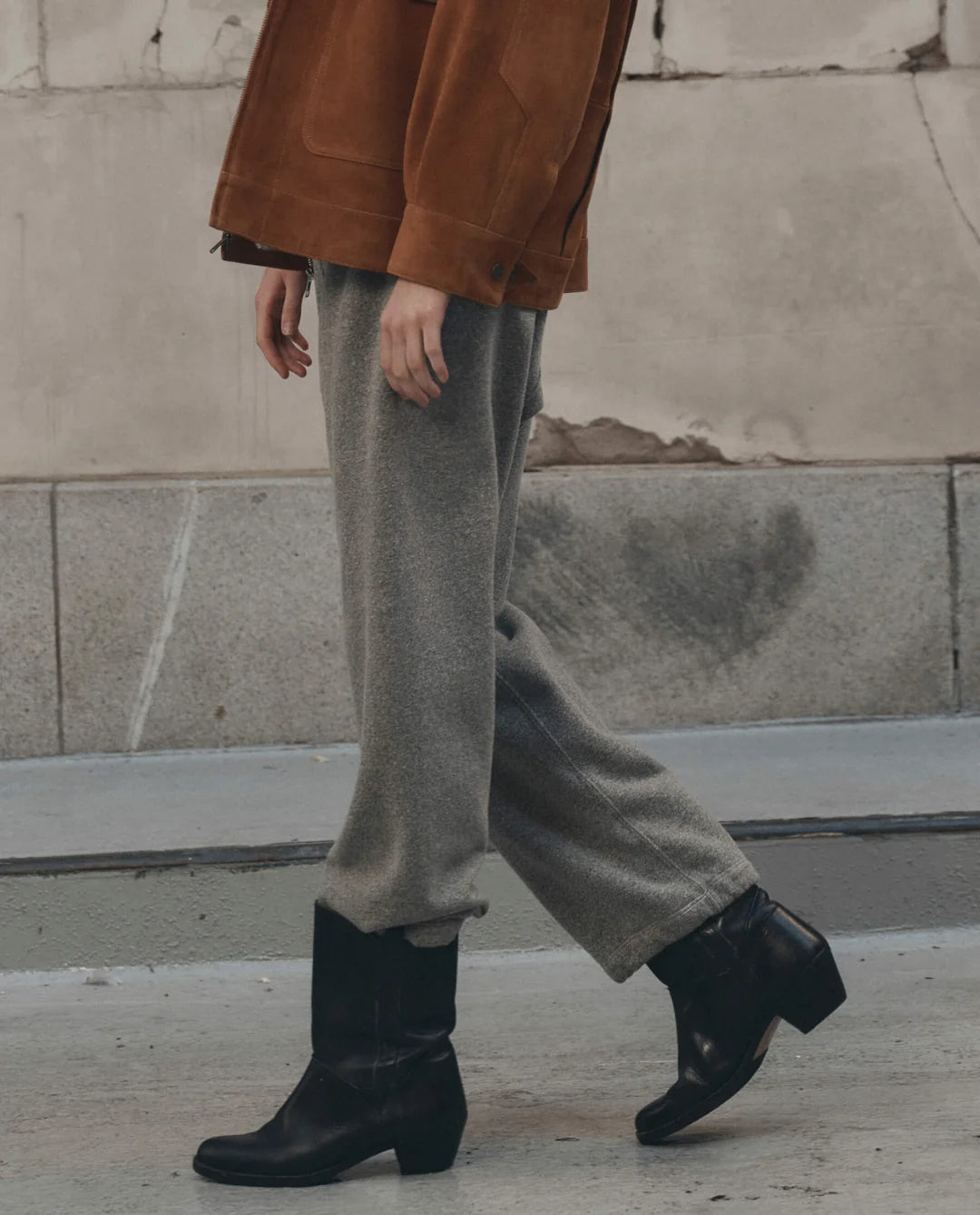 The Great Inc. Fleece Sport Sweatpant styled with black boots and a brown jacket on a city sidewalk by a weathered wall.