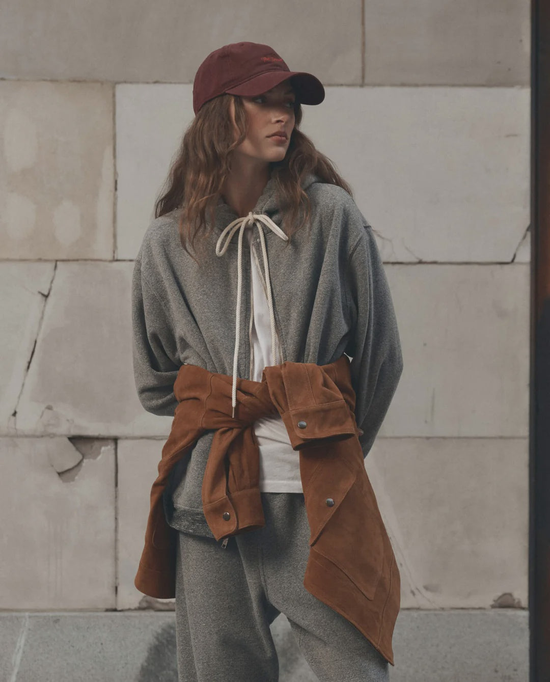 The Great Inc. Fleece Campus Hoodie in red, styled with a cap, worn casually by a textured concrete wall.
