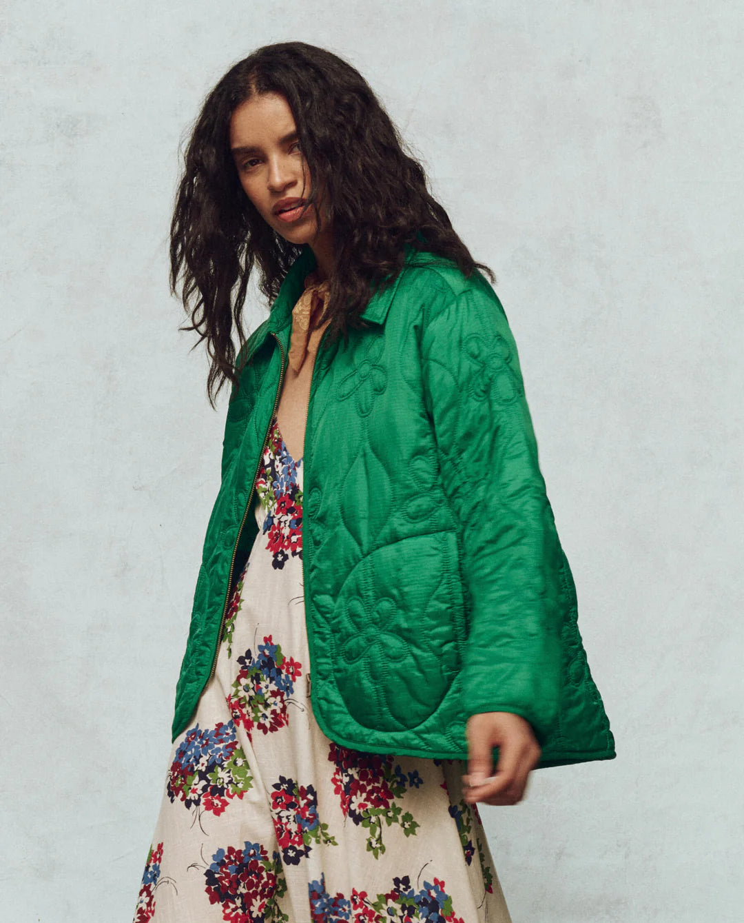 A person with long, wavy hair wears The Daisy Quilted Jacket by The Great Inc., in vibrant green featuring a daisy motif, over a floral dress adorned with red, blue, and white flowers against a muted textured off-white background.