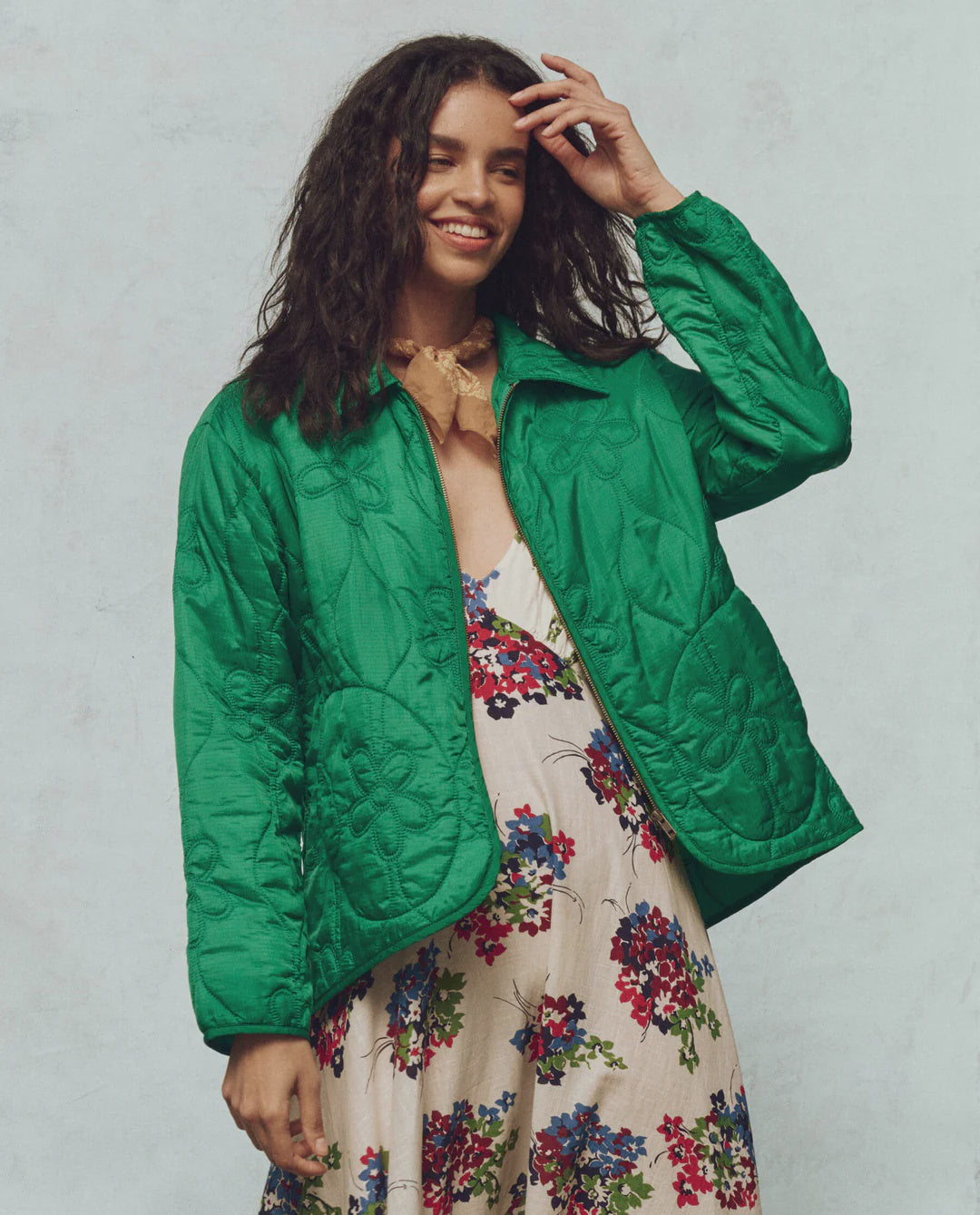 A person with long, wavy hair smiles while wearing The Daisy Quilted Jacket by The Great Inc., a vibrant green piece featuring a daisy motif and floral patterns. Underneath, they wear a multi-colored floral dress, gently resting their hand on their head against a simple background.