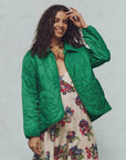 A person with long, wavy hair smiles while wearing The Daisy Quilted Jacket by The Great Inc., a vibrant green piece featuring a daisy motif and floral patterns. Underneath, they wear a multi-colored floral dress, gently resting their hand on their head against a simple background.