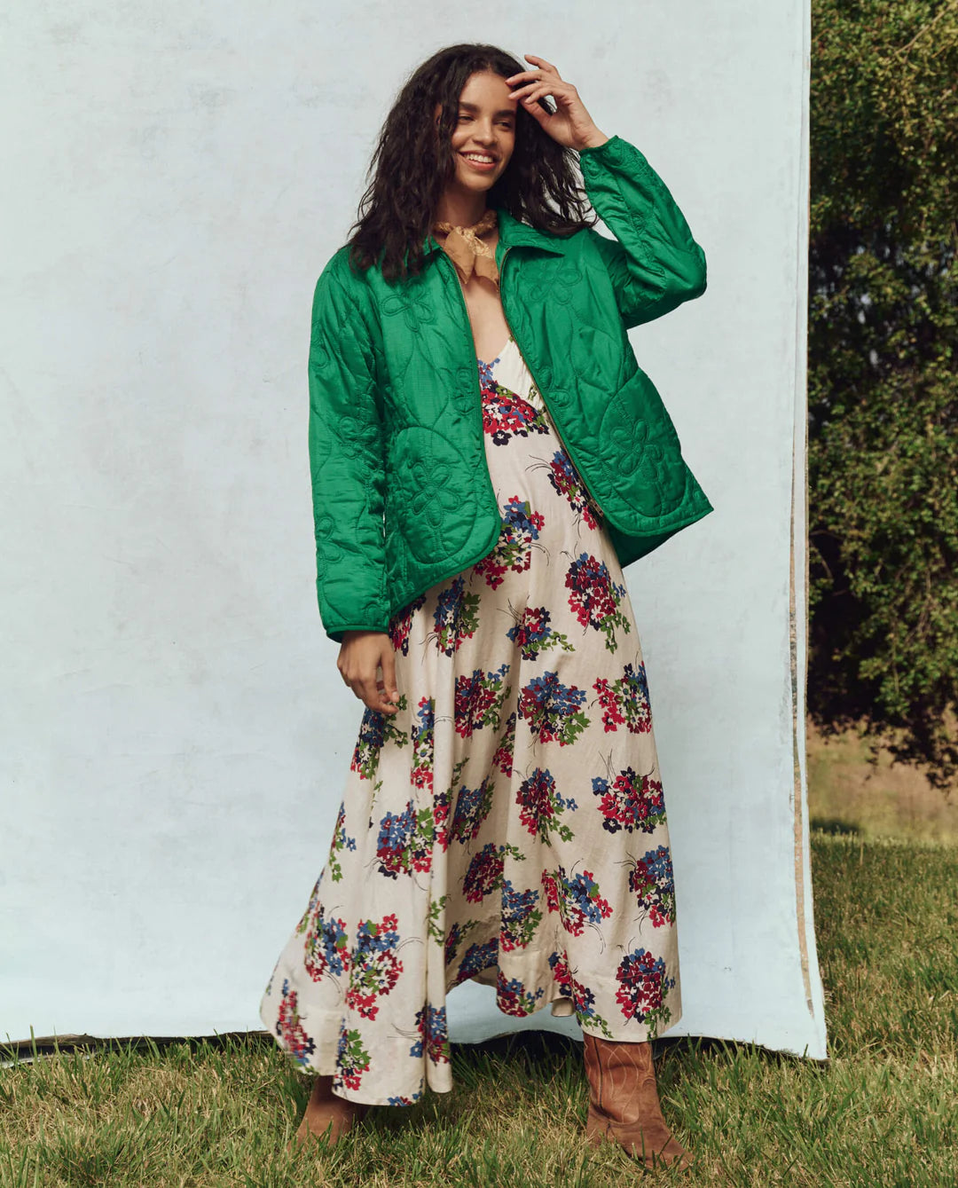 Wearing The Great Inc.'s bright green quilted jacket, featuring a daisy motif, over a long floral dress, someone stands on grass before a backdrop of trees and white. Their hand rests on their head, completing the casual look with brown boots.