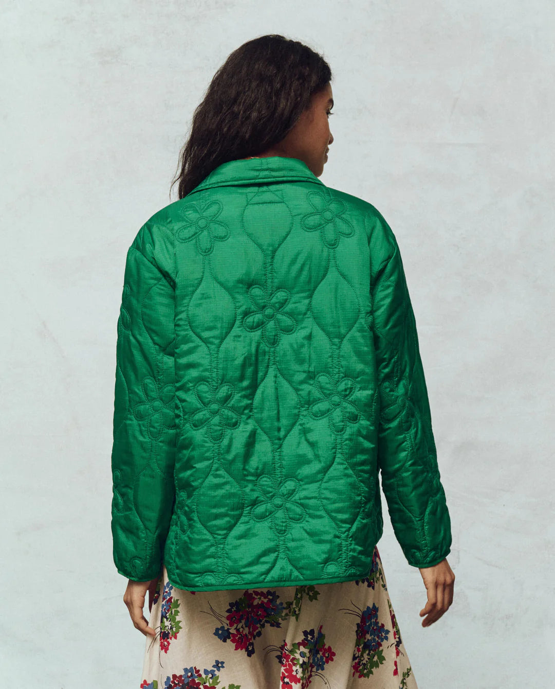 A person with long dark hair is wearing The Great Inc.'s Daisy Quilted Jacket in green, paired with a beige skirt featuring colorful floral designs, standing against a plain, light-colored wall and exuding the charm of vintage liner jackets.