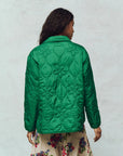 A person with long dark hair is wearing The Great Inc.'s Daisy Quilted Jacket in green, paired with a beige skirt featuring colorful floral designs, standing against a plain, light-colored wall and exuding the charm of vintage liner jackets.