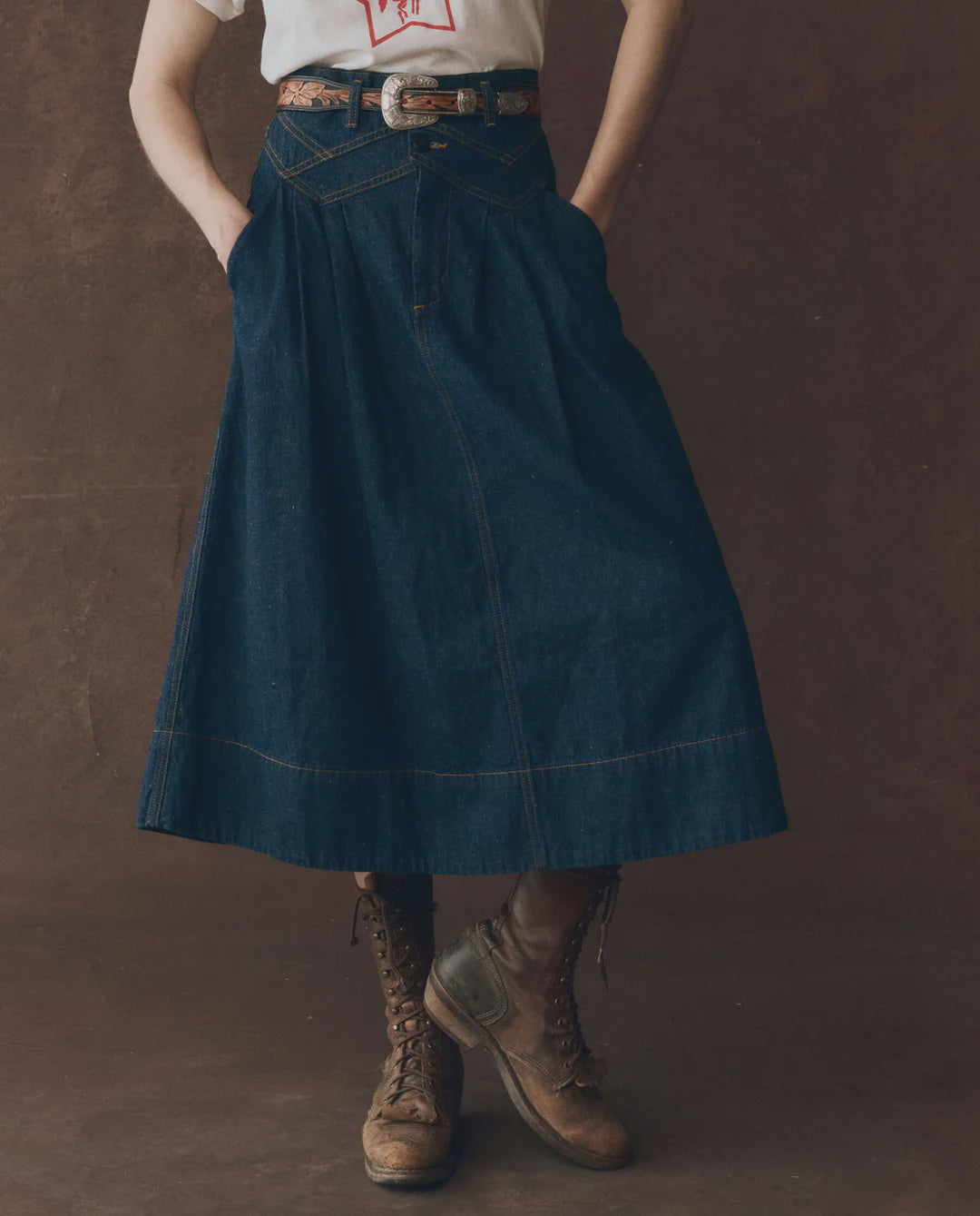 A person wearing a white T-shirt, The Great Inc.'s Field Skirt F25 in long rinse denim, a brown patterned belt, and brown lace-up combat boots stands with hands in pockets against a brown background.