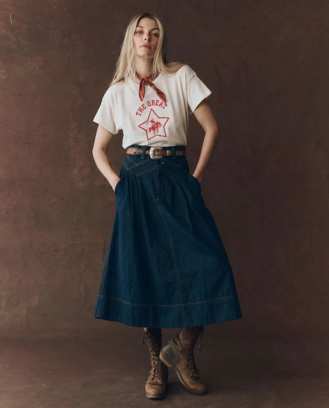 A woman stands against a brown background, wearing The Great Inc.'s The Boxy Crew 100% cotton graphic T-shirt, paired with a red bandana, high-waisted dark denim midi skirt, belt, and brown lace-up boots. Her hands rest in her skirt pockets.