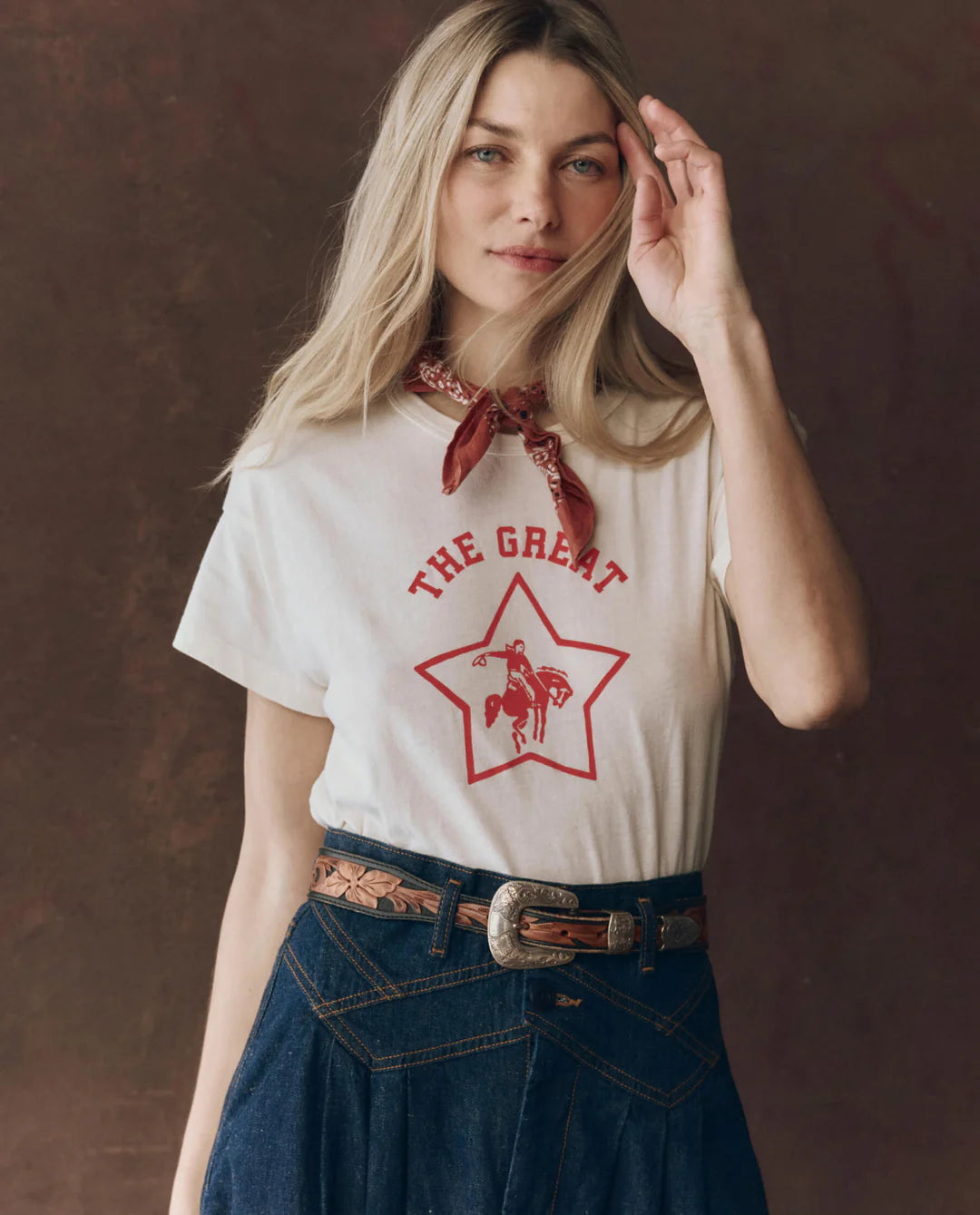 A woman with long blonde hair wears The Boxy Crew t-shirt by The Great Inc., featuring a red star and cowboy graphic, styled with a red bandana, high-waisted blue jeans, and a decorative belt, posing before a brown background.