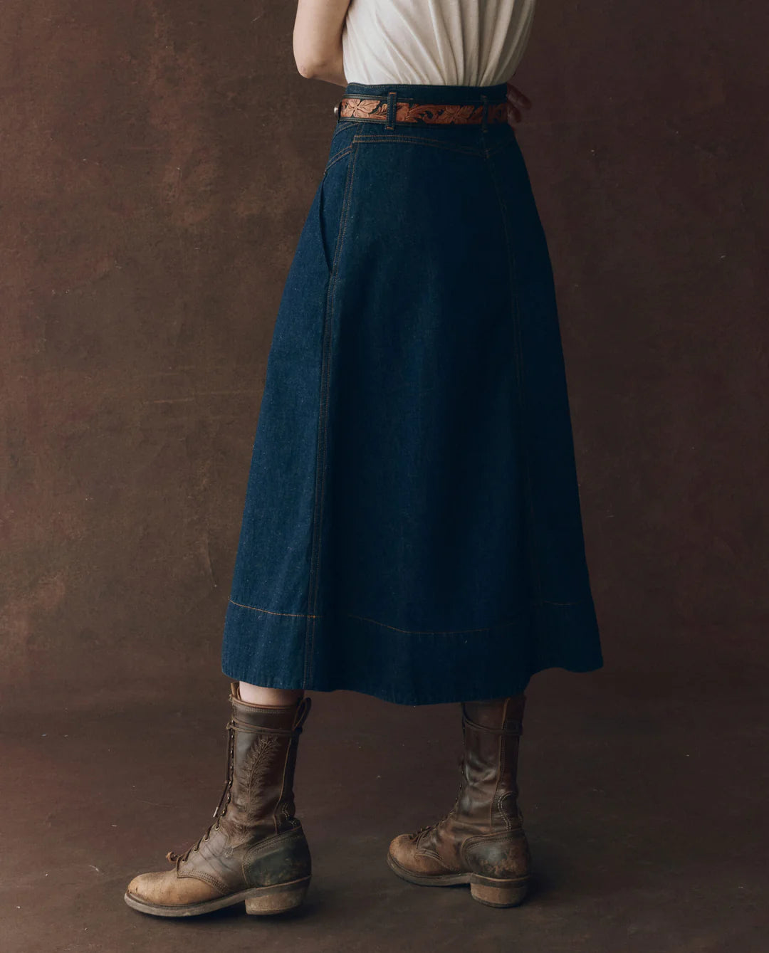 A person stands against a brown background wearing The Great Inc.’s The Field Skirt F25, styled with a white shirt and worn brown lace-up boots. Their upper body is partially out of frame.