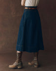 A person stands against a brown background wearing The Great Inc.’s The Field Skirt F25, styled with a white shirt and worn brown lace-up boots. Their upper body is partially out of frame.