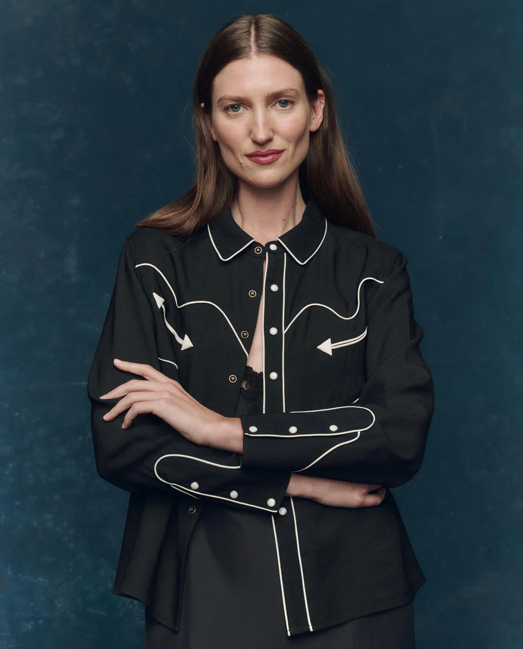 A woman with long brown hair stands with arms crossed and a slight smile, wearing The Great Inc.’s The Western Shirt—a black shirt with white piping, arrow designs, and pearl snaps—against a dark blue background.
