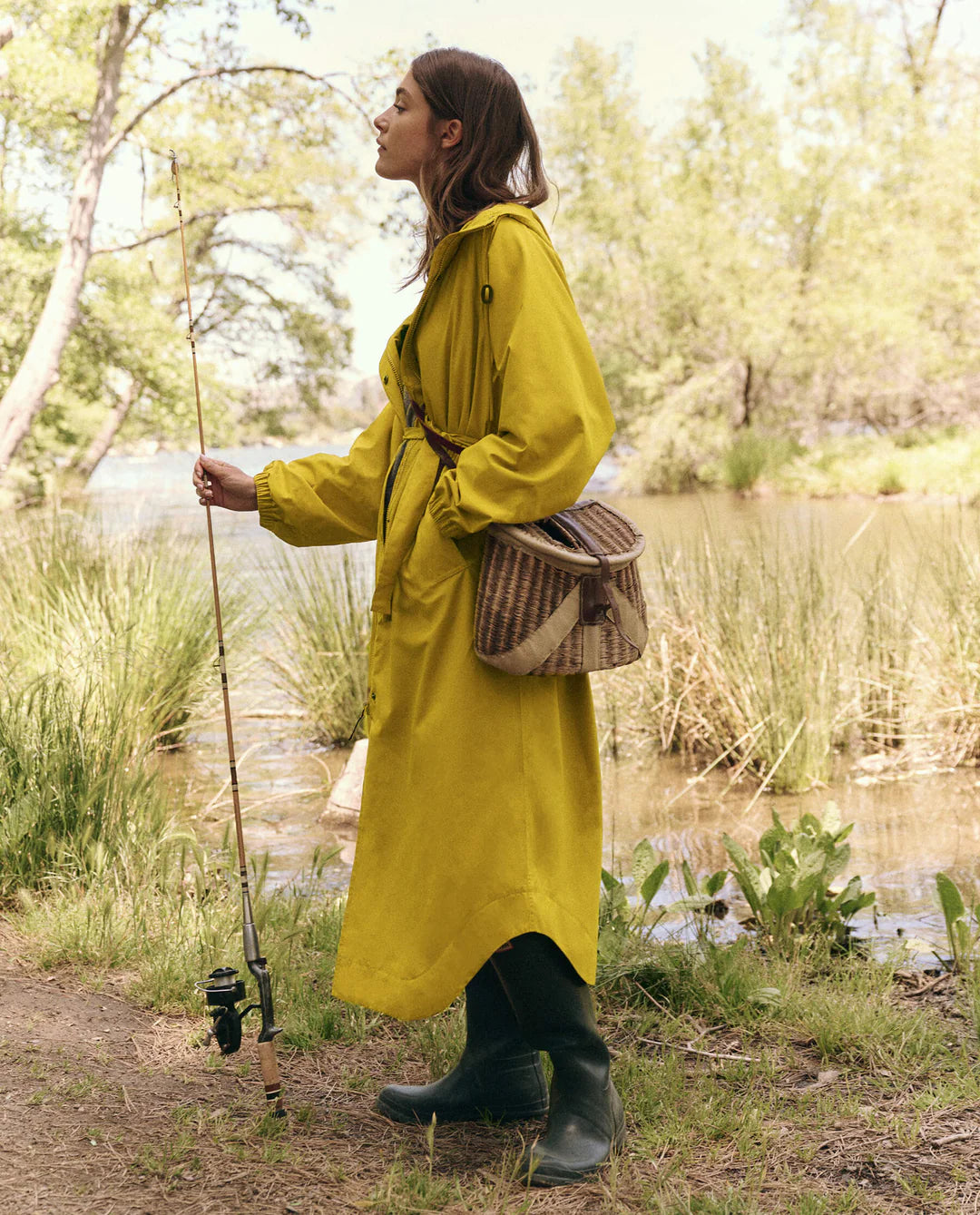 A woman wearing The Rain Trench by The Great Inc. stands by a lake holding a fishing rod. Her long hair falls over her black boots, and a brown woven satchel is slung across her body amidst trees and greenery, creating a serene scene.