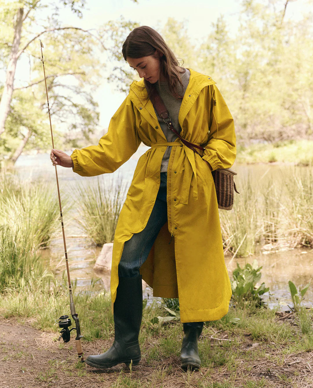 A person in "The Rain Trench" by The Great Inc. stands lakeside with a fishing rod, wearing jeans and boots. They gaze down at a small woven basket on their hip, surrounded by lush greenery and calm water in a serene setting.