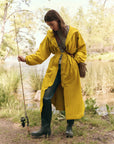 A person in "The Rain Trench" by The Great Inc. stands lakeside with a fishing rod, wearing jeans and boots. They gaze down at a small woven basket on their hip, surrounded by lush greenery and calm water in a serene setting.