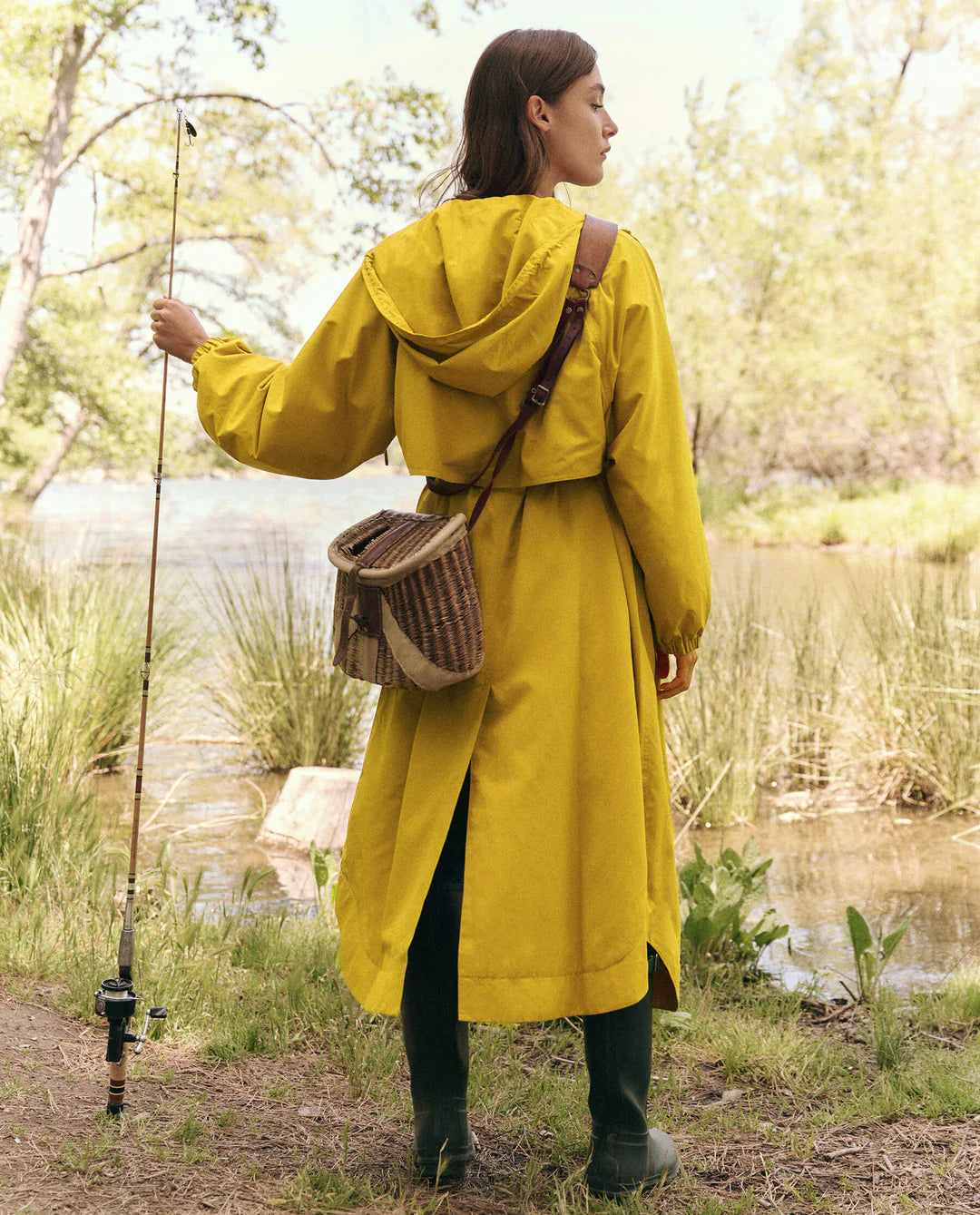 A person in The Great Inc.'s eco-friendly "The Rain Trench" hooded yellow raincoat and green boots stands by a lakeside with a fishing rod and wicker bag. Facing the water amidst trees and grass, they create a serene outdoor scene, with the recycled fabric adding an earth-conscious touch.