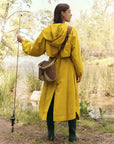 A person in The Great Inc.'s eco-friendly "The Rain Trench" hooded yellow raincoat and green boots stands by a lakeside with a fishing rod and wicker bag. Facing the water amidst trees and grass, they create a serene outdoor scene, with the recycled fabric adding an earth-conscious touch.