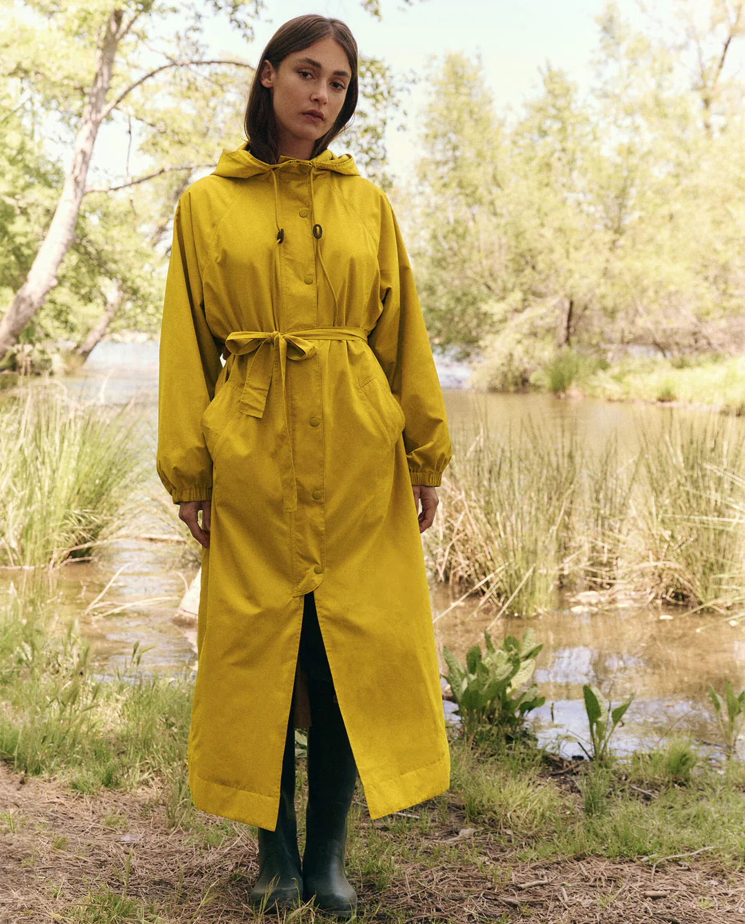 An individual wearing The Great Inc.'s Rain Trench, a hooded raincoat made from recycled materials, stands by a pond. The long belted yellow coat is paired with green boots, surrounded by grass and trees at the water's edge under a clear sky.