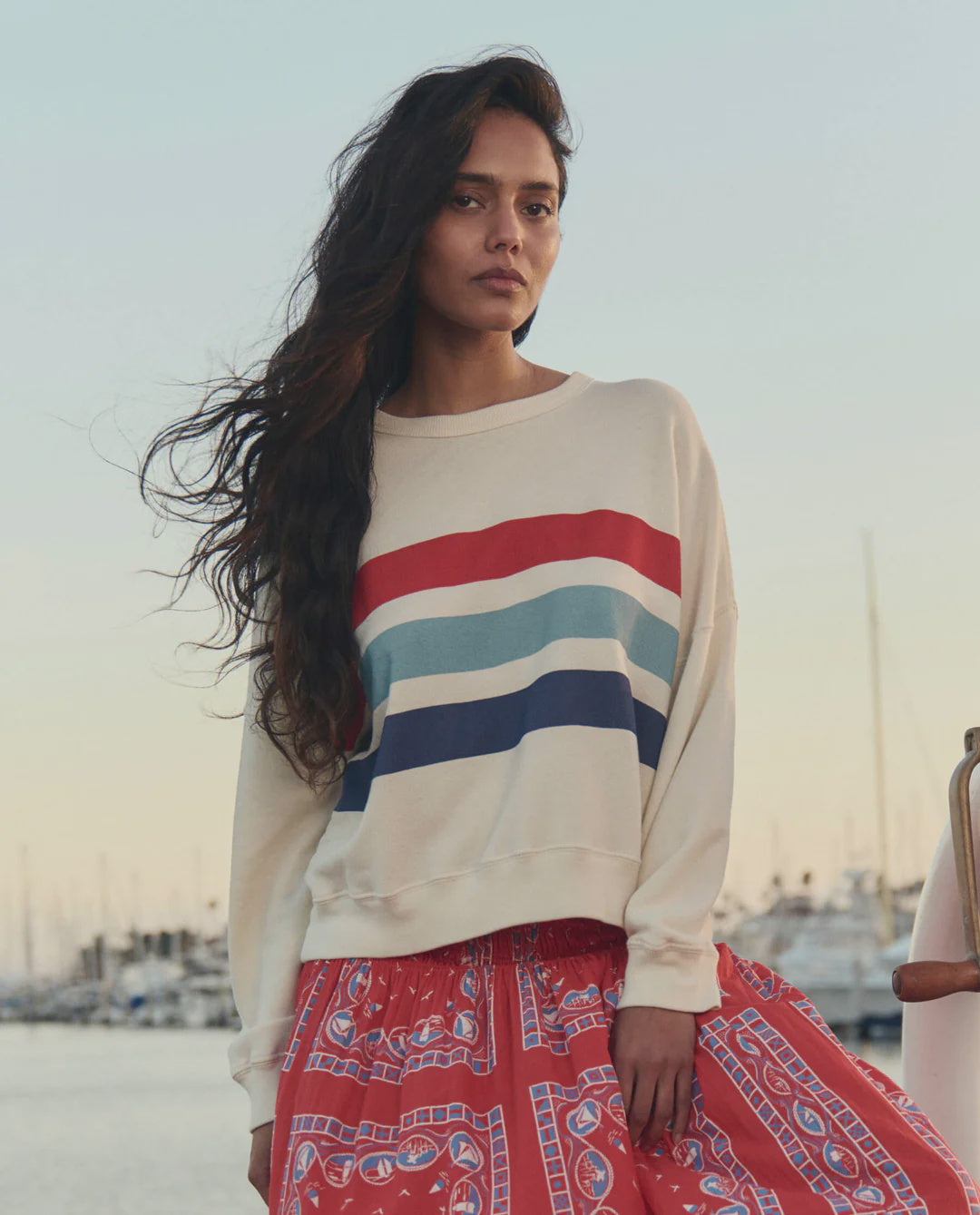 A woman with long dark hair wears The Great Inc.'s The Teammate w/saltwater Stripe sweatshirt in cream, red, teal, and navy, paired with a red patterned skirt. She stands by a marina with boats and calm water in the background.