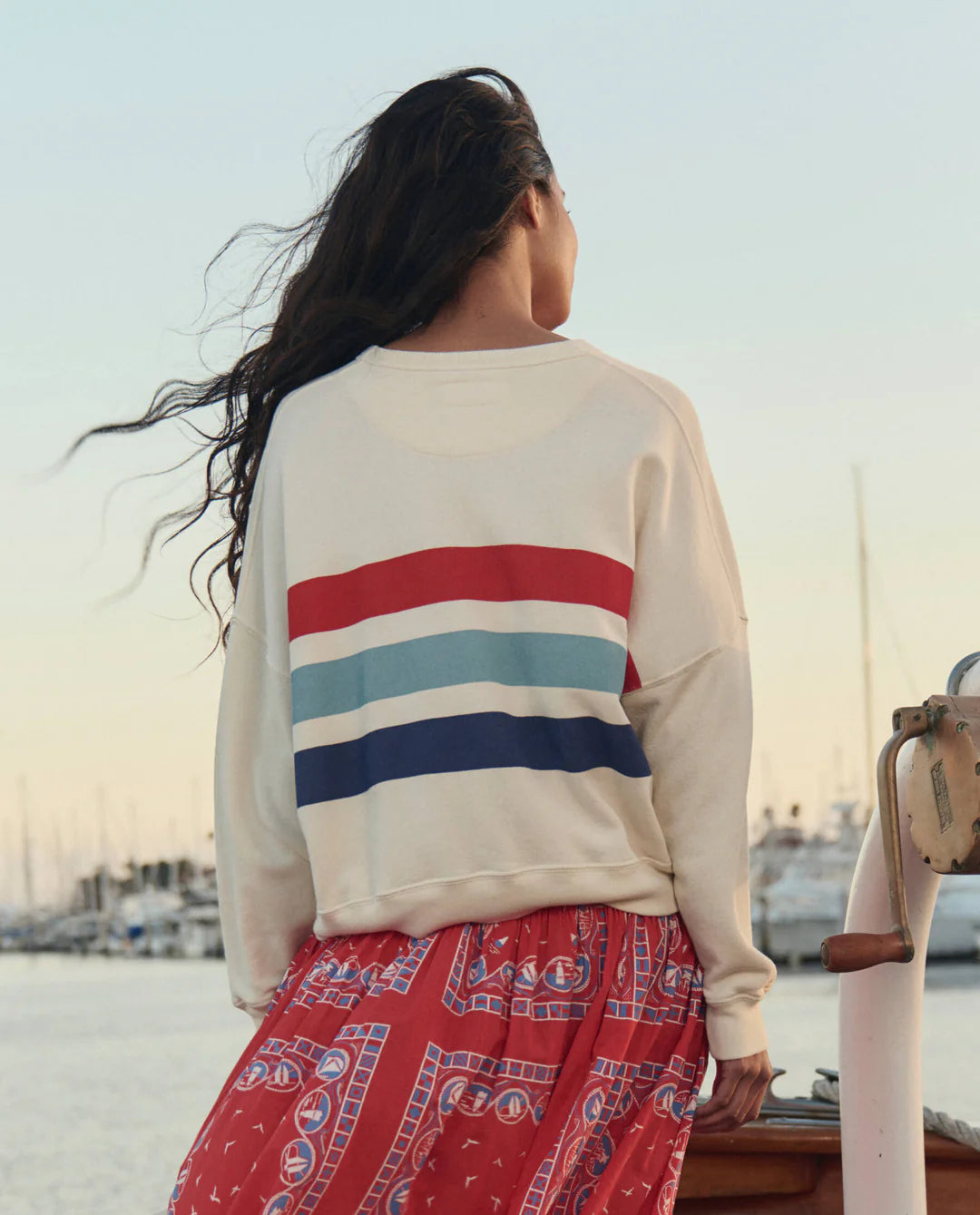 A person with long dark hair stands near a marina, facing away, wearing The Great Inc.'s The Teammate w/saltwater Stripe sweatshirt—cream with red, aqua, and navy stripes—paired with a red patterned skirt. Boats fill the background.