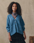 A woman with curly hair wears The Brushed Melange Cardigan by The Great Inc. in blue over a black ruffled skirt and patterned scarf, standing against a neutral beige background.