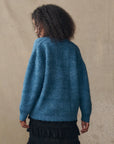 A person with curly hair, seen from behind, wears The Brushed Melange Cardigan by The Great Inc. in blue and a black ruffled skirt, standing against a neutral beige background.