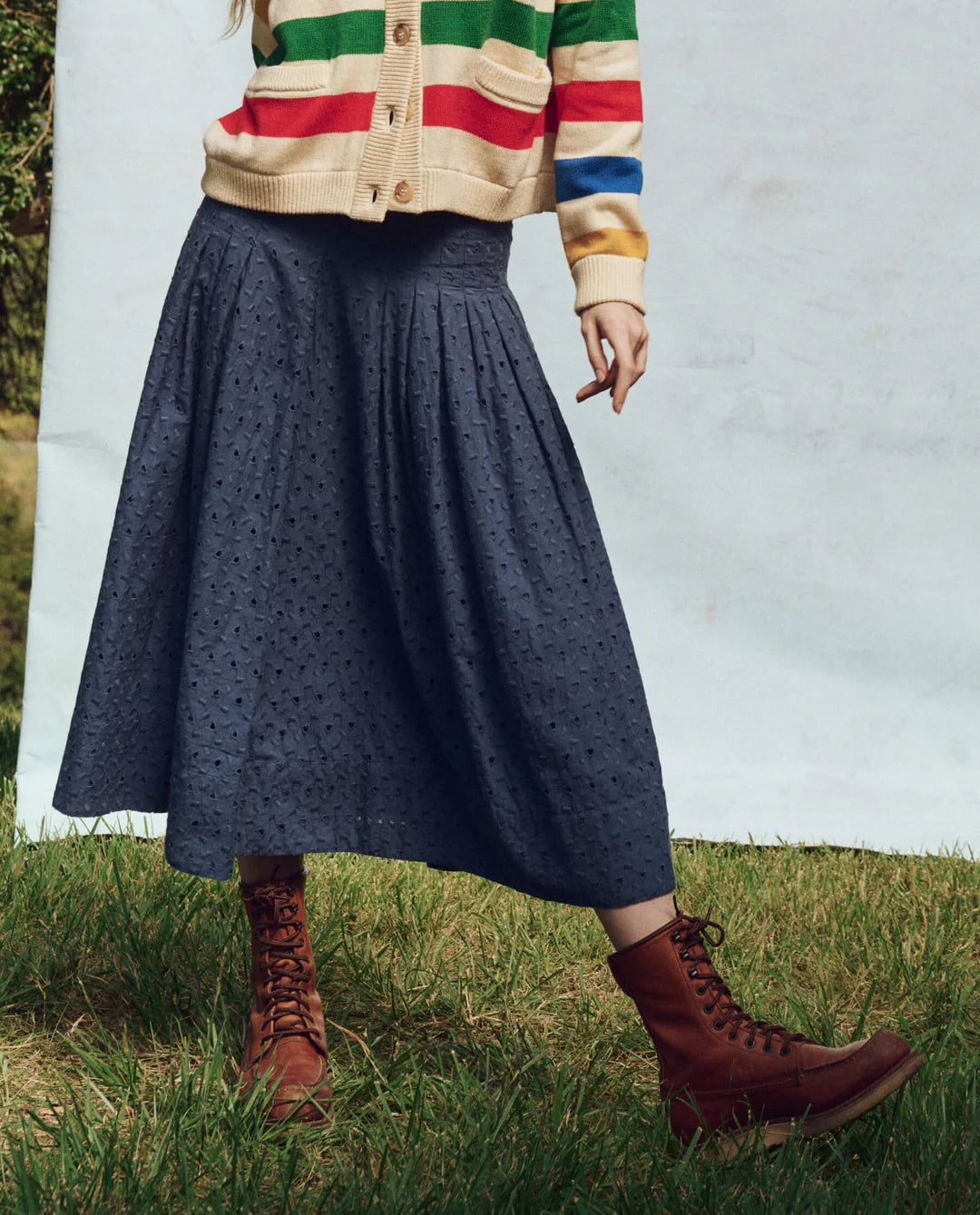 A person wearing a multicolored striped sweater, The Rhythm Skirt by The Great Inc. in dark blue, and brown lace-up boots stands on grass against a light backdrop. The person's face is not visible.