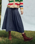 A person wearing a multicolored striped sweater, The Rhythm Skirt by The Great Inc. in dark blue, and brown lace-up boots stands on grass against a light backdrop. The person's face is not visible.