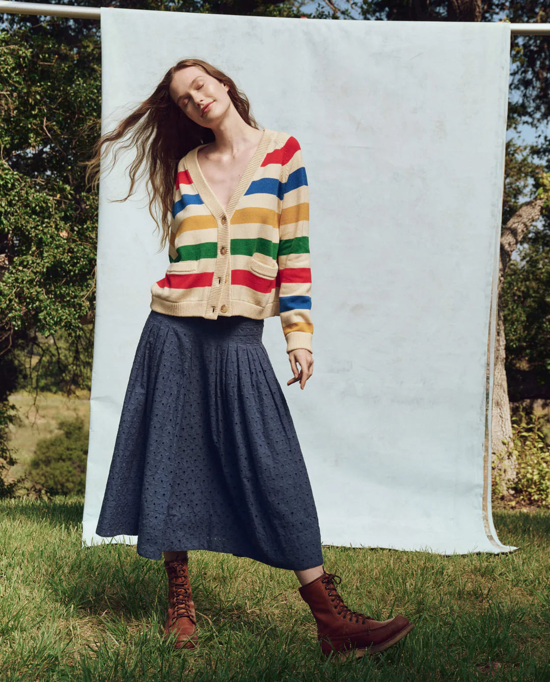 On sunny grass, a person wears a colorful striped cardigan, The Rhythm Skirt by The Great Inc. in long blue cotton eyelet style, and brown boots. They&#39;re framed by trees with a light blue backdrop, exuding Victorian charm.