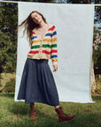 A woman with long brown hair stands on grass, wearing The Varsity Cardigan by The Great Inc., a vintage heritage-style striped piece. She pairs it with a navy midi skirt and brown boots, posing against a light blue backdrop with trees, gracing the scene with a gentle smile.