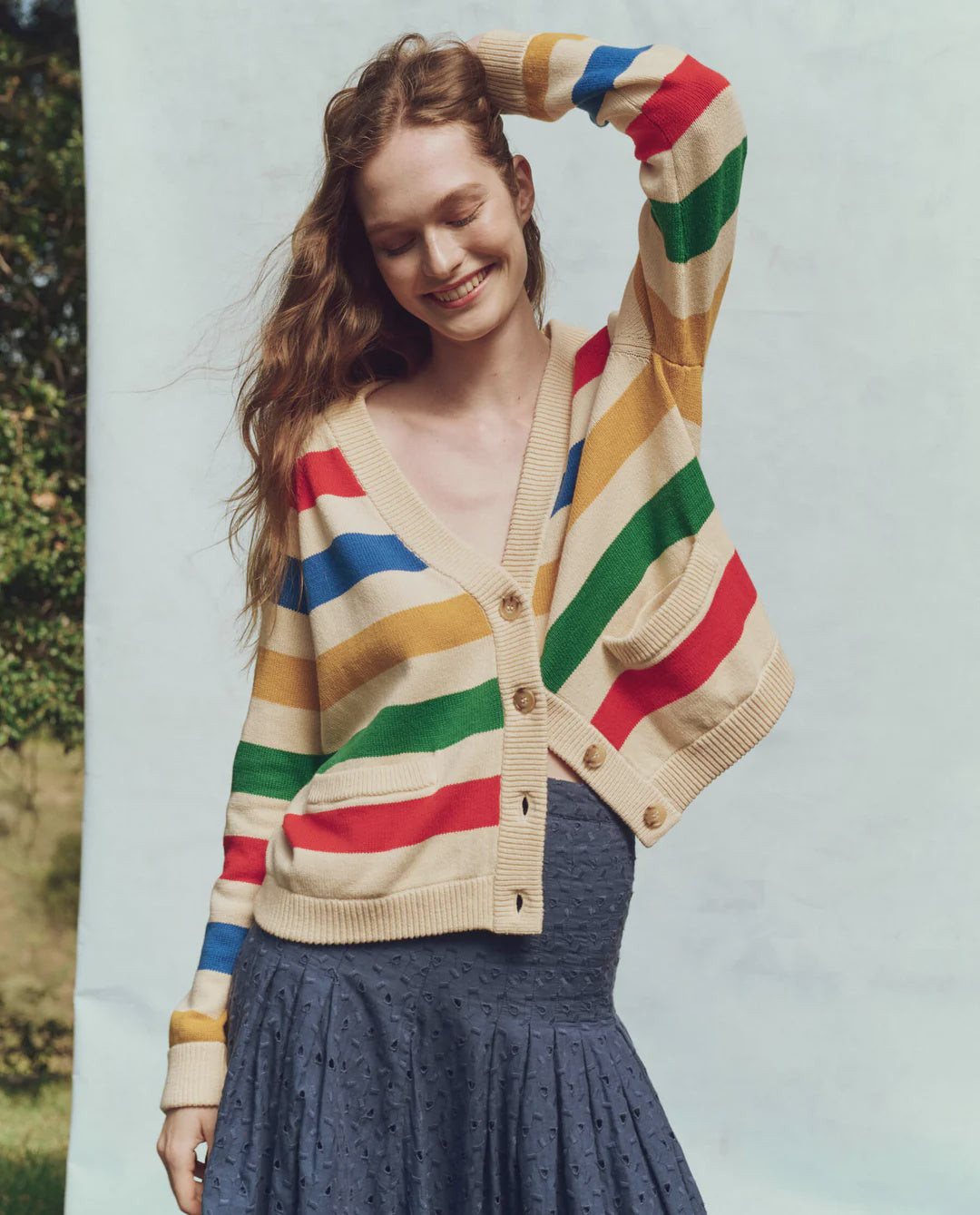 A person smiles with closed eyes, wearing The Varsity Cardigan by The Great Inc. over a dark skirt. Their right arm bends above their head against a light blue backdrop, with greenery peeking in on the left, enhancing the charm of vintage heritage styles.