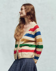 A woman with long, wavy hair stands in profile against a light background, highlighting The Great Inc.'s Varsity Cardigan featuring colorful horizontal stripes of red, blue, green, and yellow in a cozy cotton blend. A dark textured skirt completes her look.