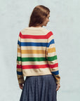 Seen from the back, an individual with long hair dons The Great Inc.'s The Varsity Cardigan, a cream cotton blend adorned with blue, red, green, and yellow stripes. A blue skirt with small polka dots on a light background completes the look.