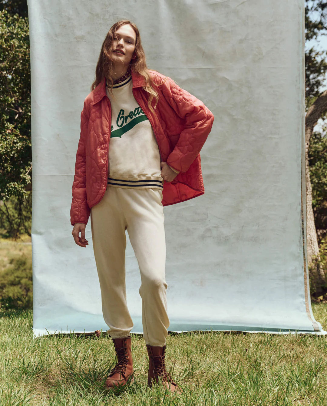 In front of a plain backdrop, someone stands on grass, wearing a red quilted jacket over The Great Inc.'s Varsity College Sweatshirt with Crest Graphic, light beige pants, and brown boots. Trees and greenery are visible in the background.