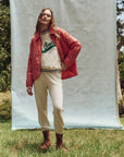 In front of a plain backdrop, someone stands on grass, wearing a red quilted jacket over The Great Inc.'s Varsity College Sweatshirt with Crest Graphic, light beige pants, and brown boots. Trees and greenery are visible in the background.